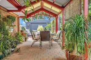 En overdækket terrasse med et bord og stole, omgivet af plantekrukker og udsigt til en bygning.