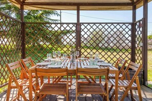 A covered outdoor dining area with wooden tables and chairs, a lattice fence, and a view of greenery.
