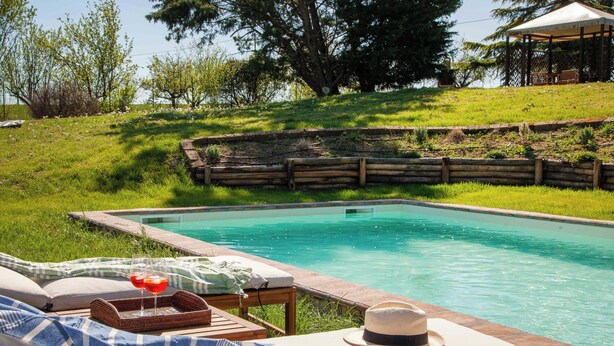 Un área junto a la piscina con una tumbona, una mesita con una bandeja y vasos, y un sombrero en la silla.