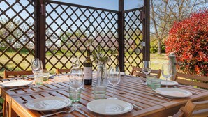 Una mesa de madera dispuesta para comer, con copas de vino, platos, cubiertos, una botella y un florero con flores.