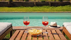 Ett träbord med en glasskål full av chips och två glas med röd dryck, med utsikt över en pool med ett trästaket och grönska i bakgrunden.