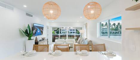 A modern dining area with a white table set for four, two pendant lights, and a view of a balcony with a pool.