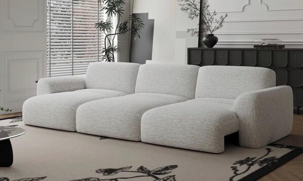 Un canapé de couleur claire dans un salon moderne, agrémenté d’un tapis à motifs, d’une plante et d’un meuble de rangement pour appareils électroniques aux tons sombres.