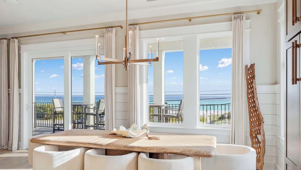 A spacious dining area with a wooden table and white chairs, large windows with a view of the sea, and a balcony with outdoor seating. - NEW Laguna Serenity | 7BR Gulf Front | Pool | LSV/Bikes (Panama City Beach)