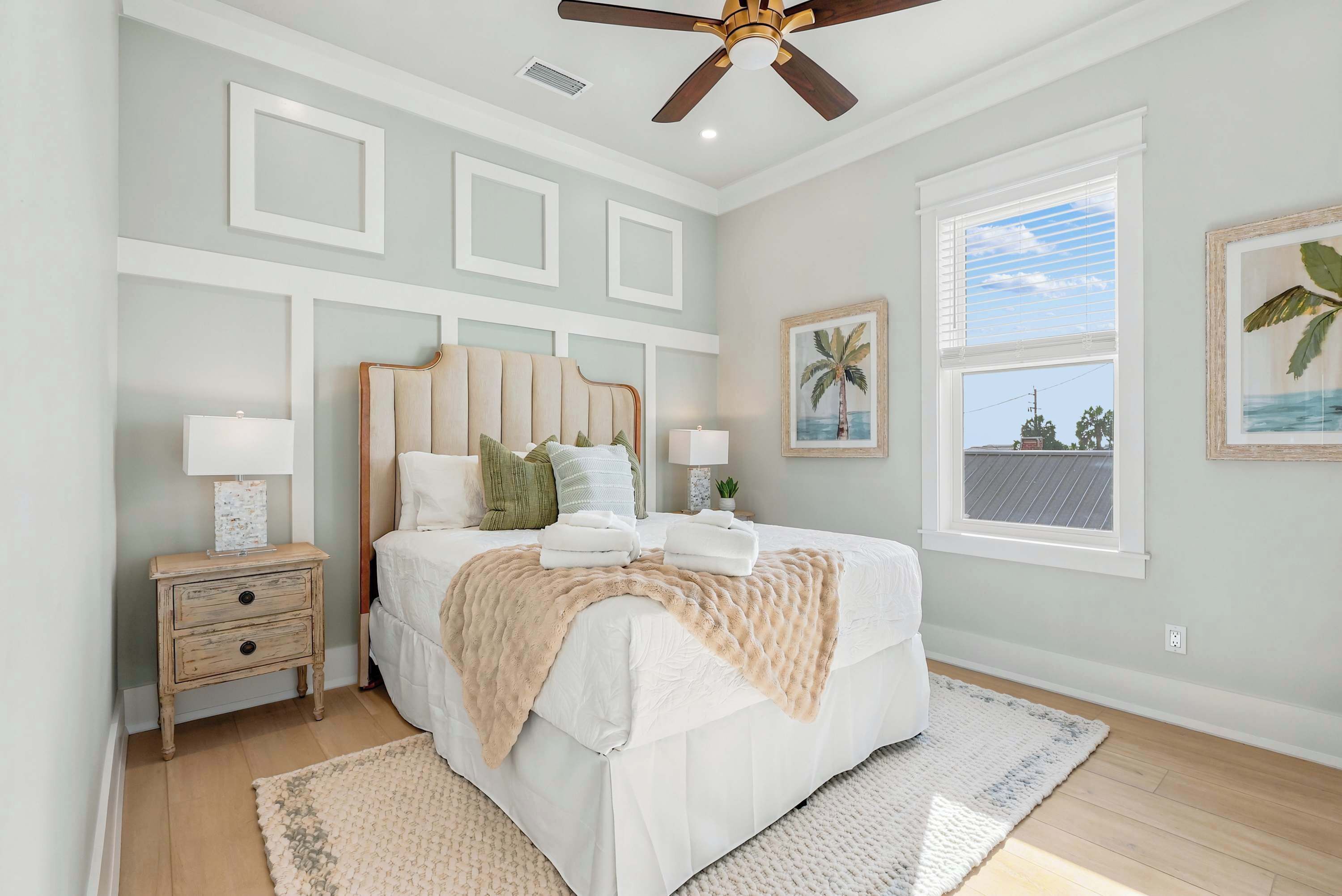 A bedroom with a bed, bedside tables, a ceiling fan, a window with a view, and wall-mounted artwork.