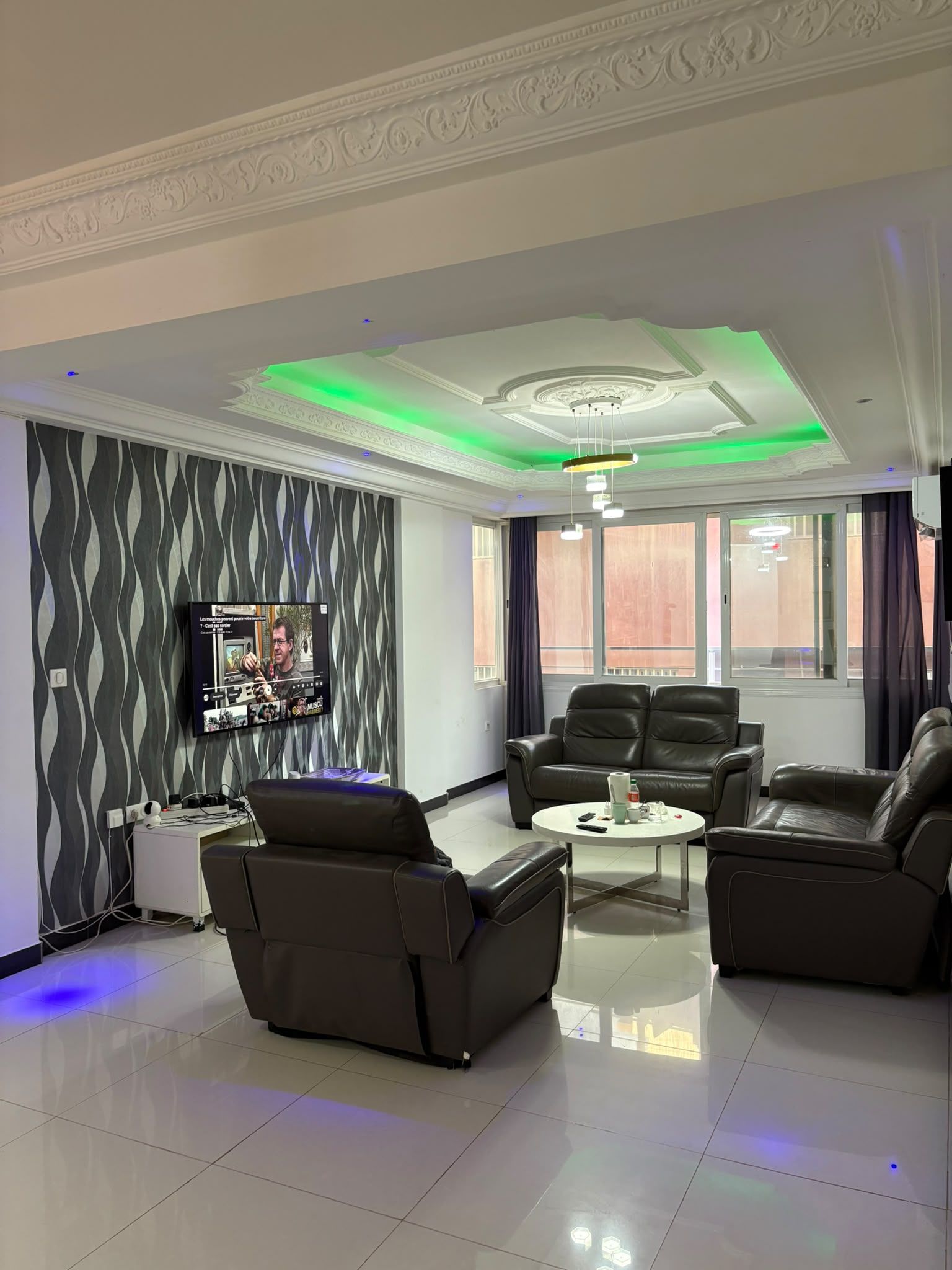 A modern living room with a flat-screen TV, leather sofas, a coffee table, and a patterned wallpaper.