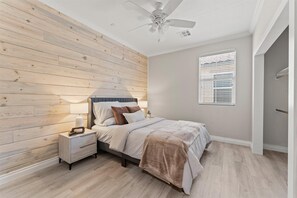 Ein Schlafzimmer mit Bett, Nachttisch, Lampe, Deckenventilator und einem Fenster mit Jalousien.