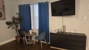 Une pièce dotée d’un téléviseur à écran plat fixé au mur, d’une petite table ronde avec deux chaises, d’une plante en pot et d’une commode de couleur sombre.