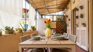A covered patio with a table set for dining, surrounded by potted plants and a decorative vase.