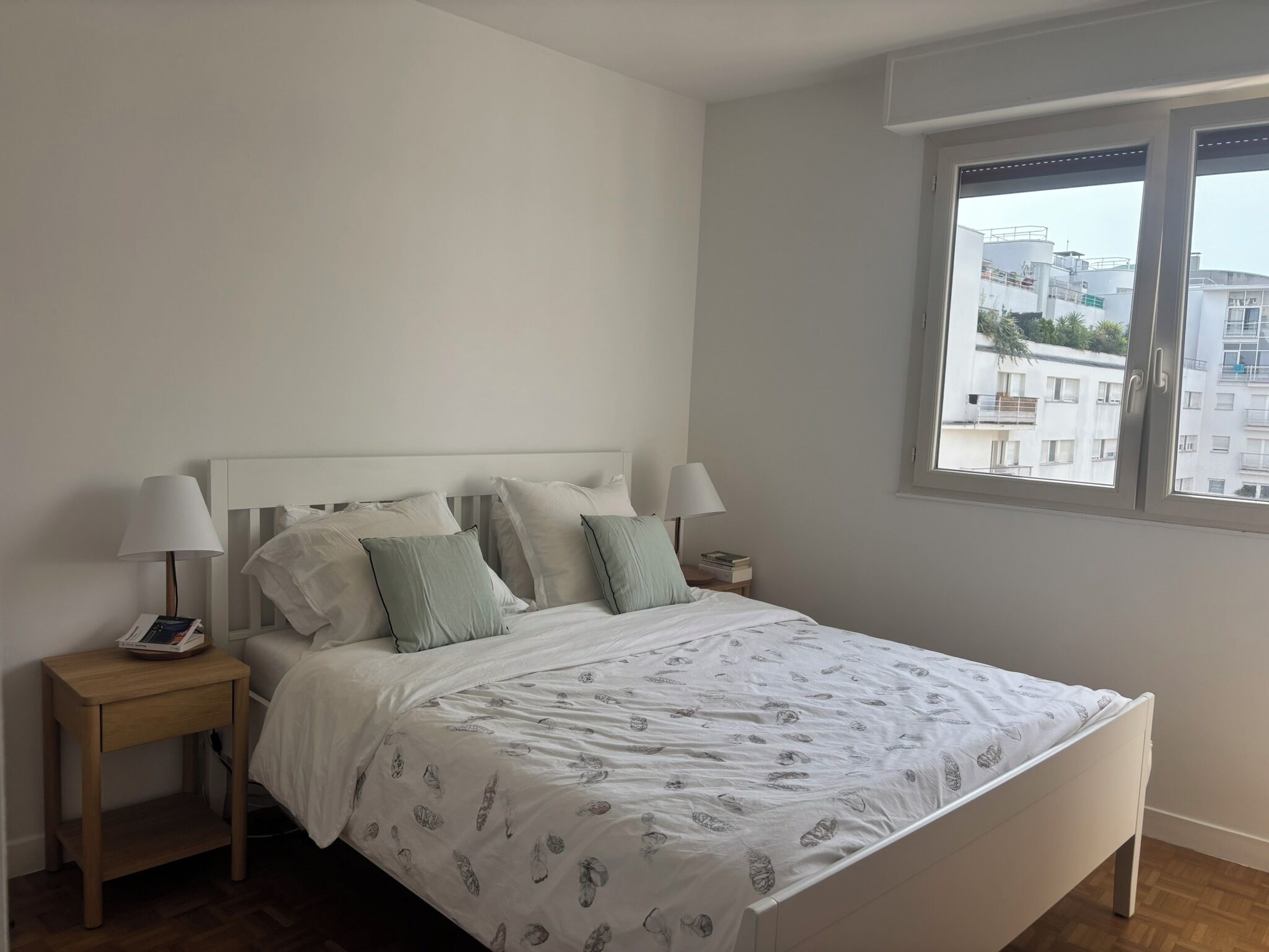 A bedroom with a bed, two bedside tables, a window, and a view of buildings outside.