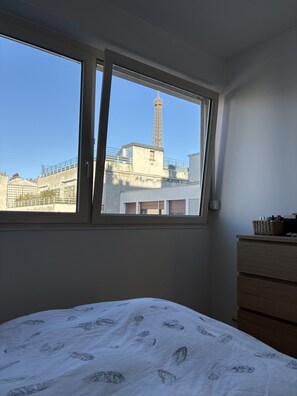 Ein Schlafzimmer mit großem Fenster, einem Bett mit blattbedeckter Bettdecke, einer Holzkommode und Blick auf einen Turm.