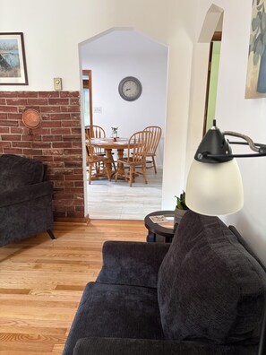 A living room with a dark sofa, a lamp, a dining area with a wooden table and chairs, and a brick wall.