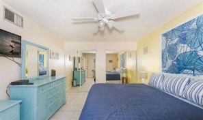 Une chambre à coucher comprenant un lit, une commode, un ventilateur de plafond, un tableau accroché au mur et une porte donnant sur une autre pièce.