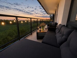 Ein Balkon mit einer Couch, einem Tisch mit einer Vase und Weingläsern sowie Blick auf ein Feld und einen Sonnenuntergang.