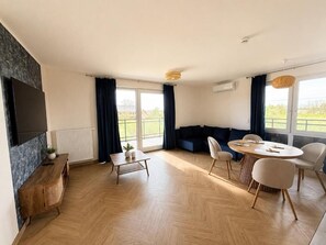 Ein modernes Wohnzimmer mit Sofa, Esstisch mit Stühlen, einem an der Wand befestigten Fernseher und großen Fenstern.