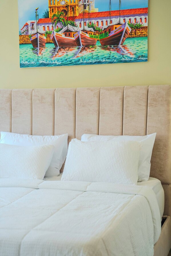 A bed with white bedding and a headboard, a painting of boats and a building on the wall behind it.