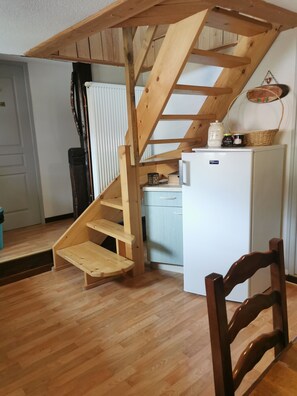 Una escalera de madera que lleva a una zona de entrepiso, un refrigerador blanco y una mesa de comedor de madera con sillas.