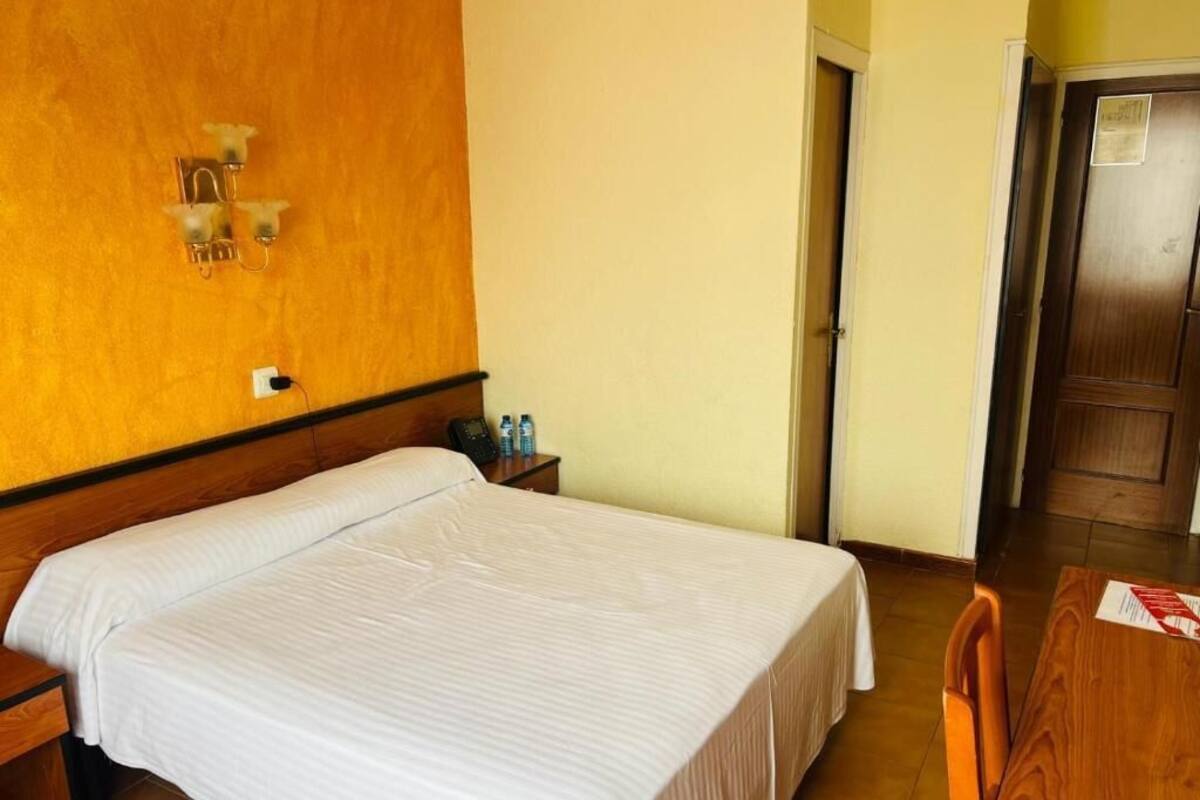 Una habitación de hotel con cama, mesita de noche, lámpara y una puerta de madera.