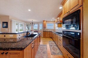 A kitchen with wooden cabinets, a granite countertop, a stainless steel oven, and a large window. - #HabitueHomes - The Quail Ridge Overlook (Boise)