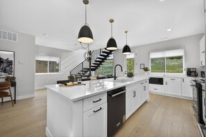 A modern kitchen with a central island, black pendant lights, white cabinetry, and a staircase leading to an upper level. - Fresh Tracks @ Peak View Ridge (Winter Park)