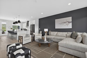 Ein modernes Wohnzimmer mit einem grauen Sofa, einem runden Couchtisch und einer Küchenzeile mit weißen Schränken und schwarzen Pendelleuchten.