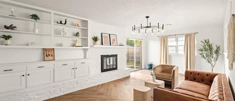 A modern living room with a white brick fireplace, wooden flooring, a brown sofa, and a white shelving unit with various decorative items.