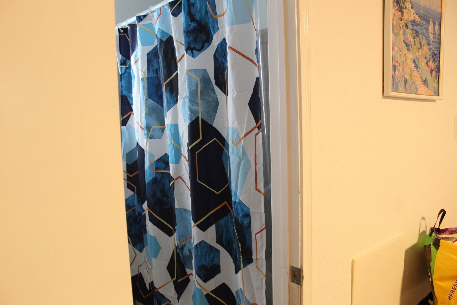 Een badkamer met een douchegordijn met een geometrisch patroon, een schilderij aan de muur en een gele tas op de vloer.