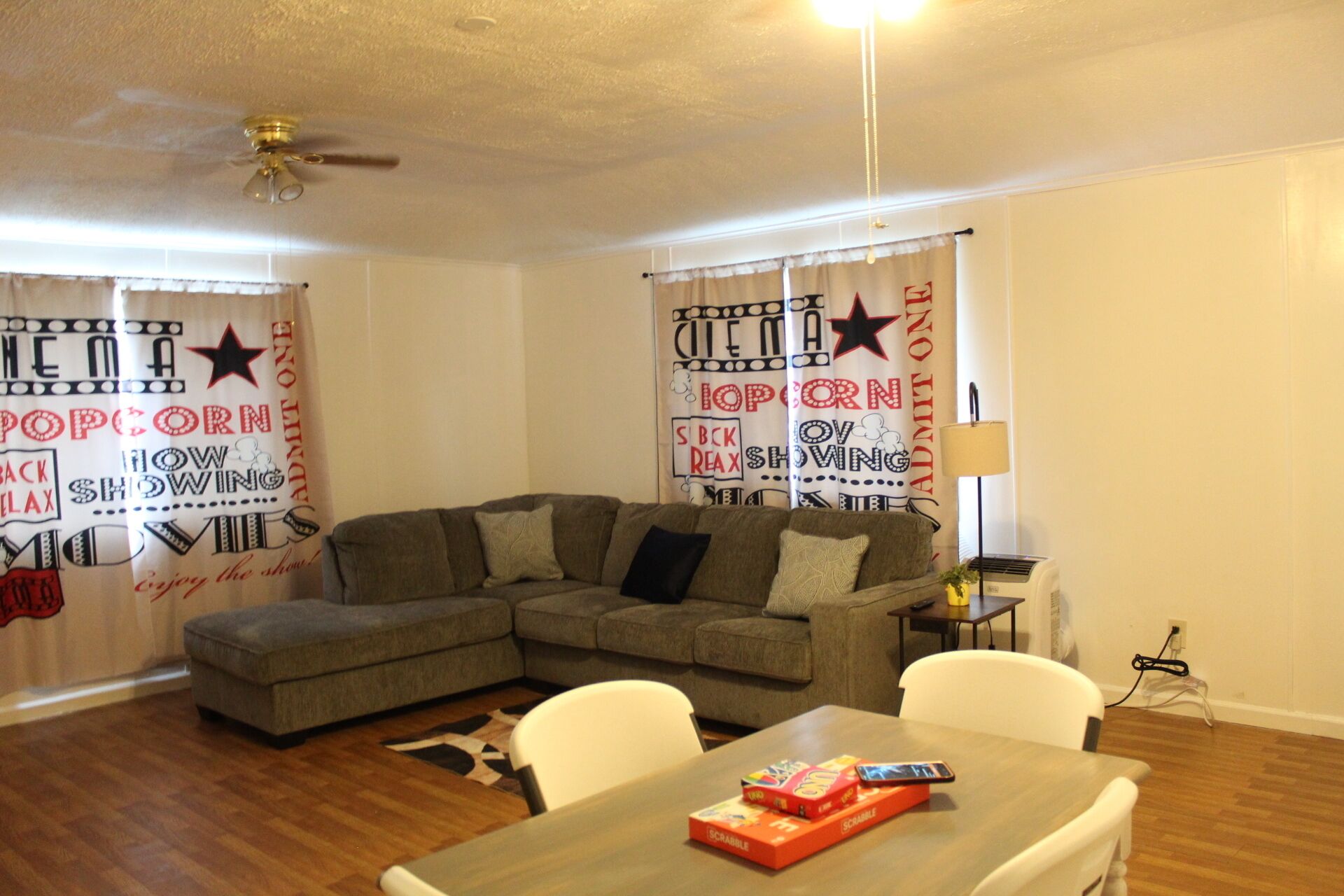 A living room with a sectional sofa, a dining table with Scrabble tiles, and curtains with cinema-themed designs.
