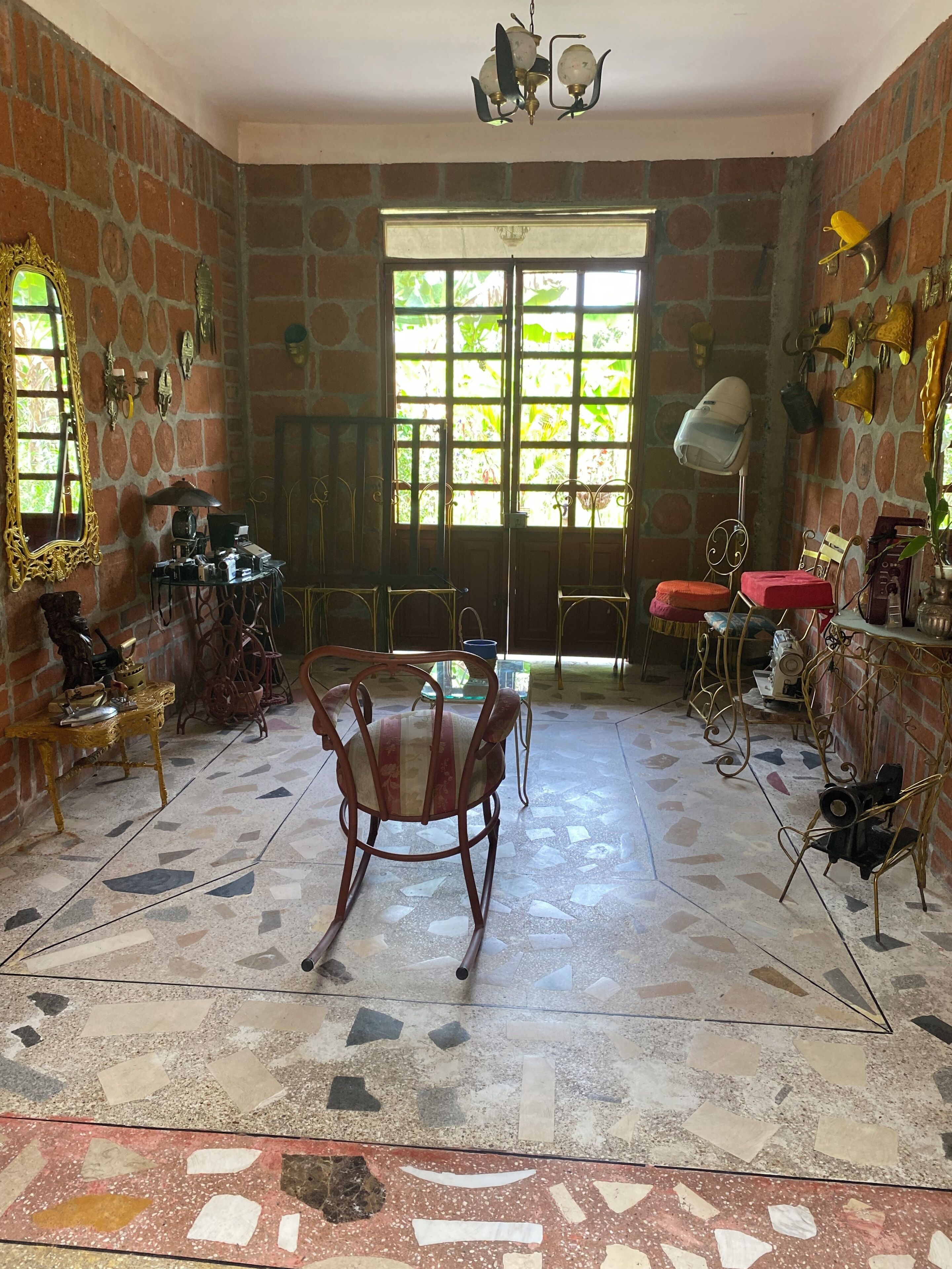 Une pièce avec un sol carrelé, un fauteuil à bascule en bois, une table, une fenêtre donnant sur un paysage verdoyant et un mur en briques.