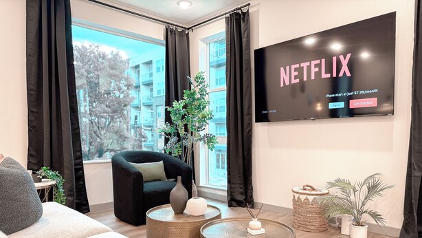 A modern living room with a large TV displaying a Netflix advertisement, a comfortable sofa, a black armchair, and a view of a building through large windows. - Dallas Downtown, FIFA Close to All the FUN! (Dallas)