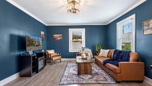A living room with a blue wall, a wooden coffee table, a brown sofa, a TV, and a window with blinds. - The Retreat at Harpers Ferry (Bolivar)