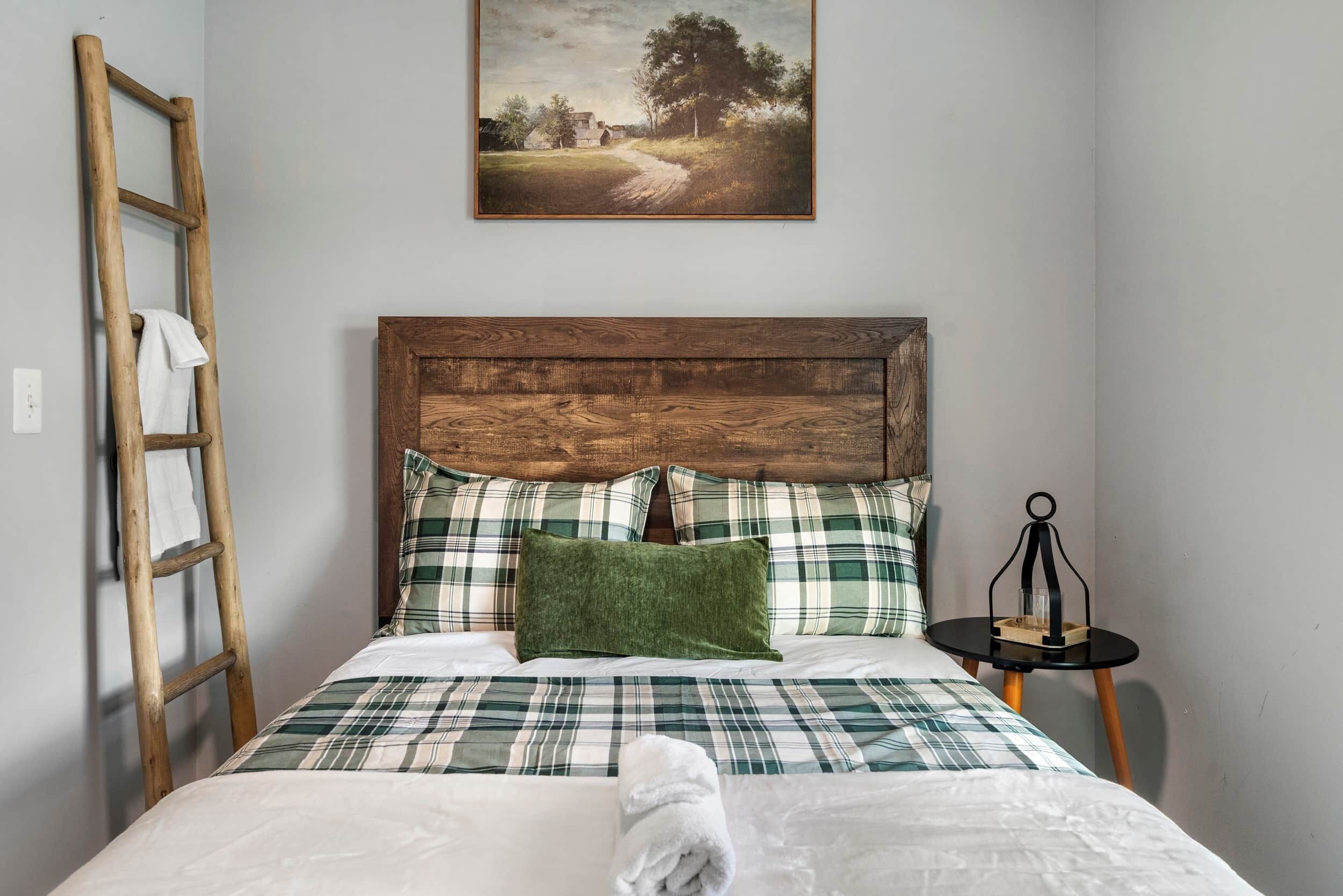 Ein Bett mit Karomuster-Bettwäsche, ein Holz-Kopfteil, ein grünes Kopfkissen und eine Lehne an der Wand. Darüber hängt ein Bild.