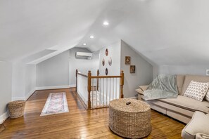 Una habitación estilo loft con piso de madera, un sofá, un reposapiés tejido, una escalera y una barandilla.
