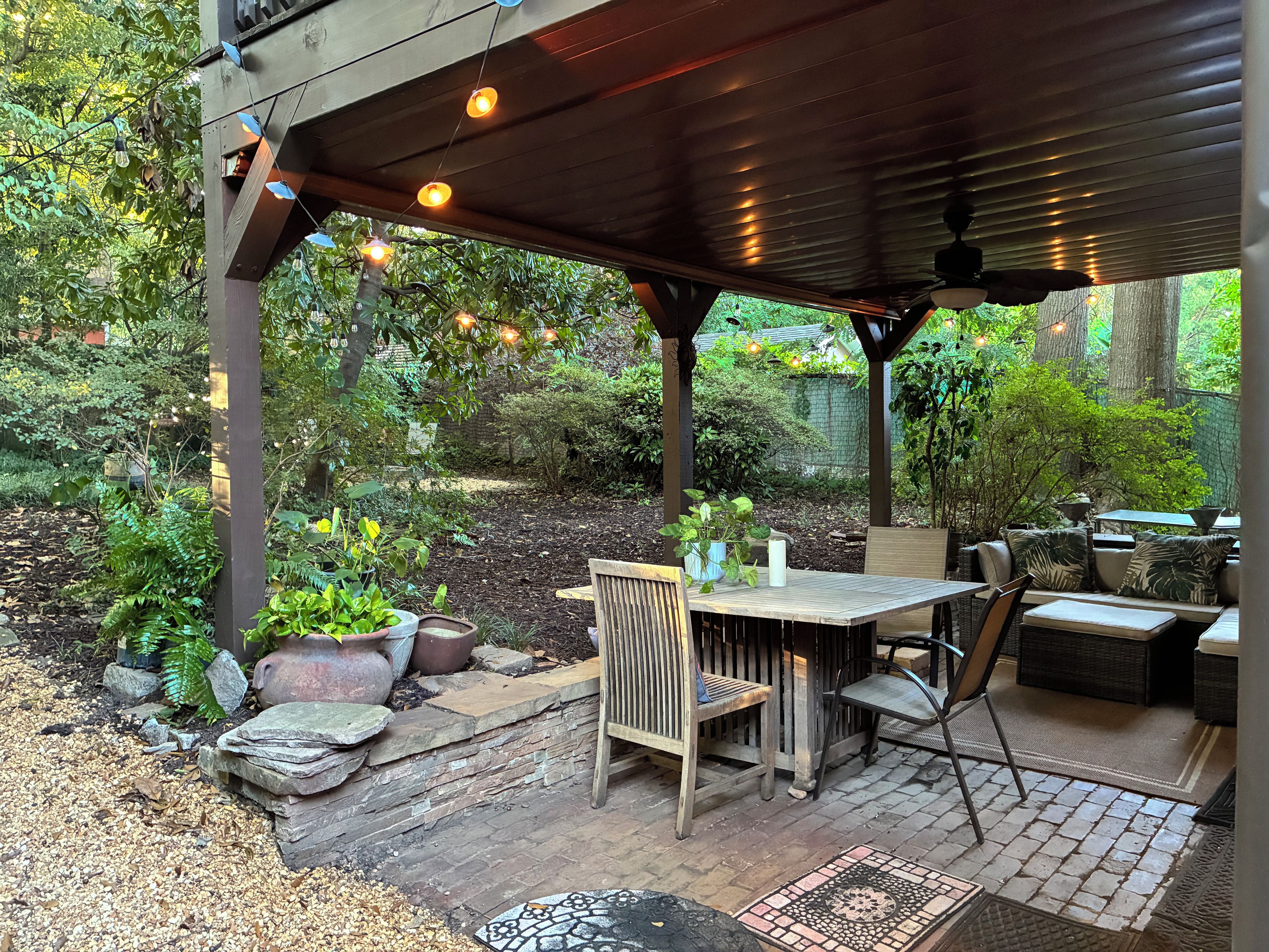 Un patio al aire libre con mesa y sillas de madera, rodeado de plantas en macetas y un sendero de ladrillo.