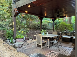 Une terrasse extérieure avec une table et des chaises en bois, entourée de plantes en pot et bordée d’un chemin en briques.