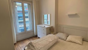 Ett sovrum med en stor säng, en byrå, en spegel och ett fönster med utsikt över balkonger.