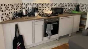 Ett kompakt kök med mikrovågsugn, spis och ugn, en liten arbetsbänk och en grå soffa i vardagsrummet.