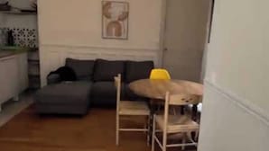 Ett vardagsrum med en soffa, ett matbord med stolar, en köksdel med skåp och en dörr.