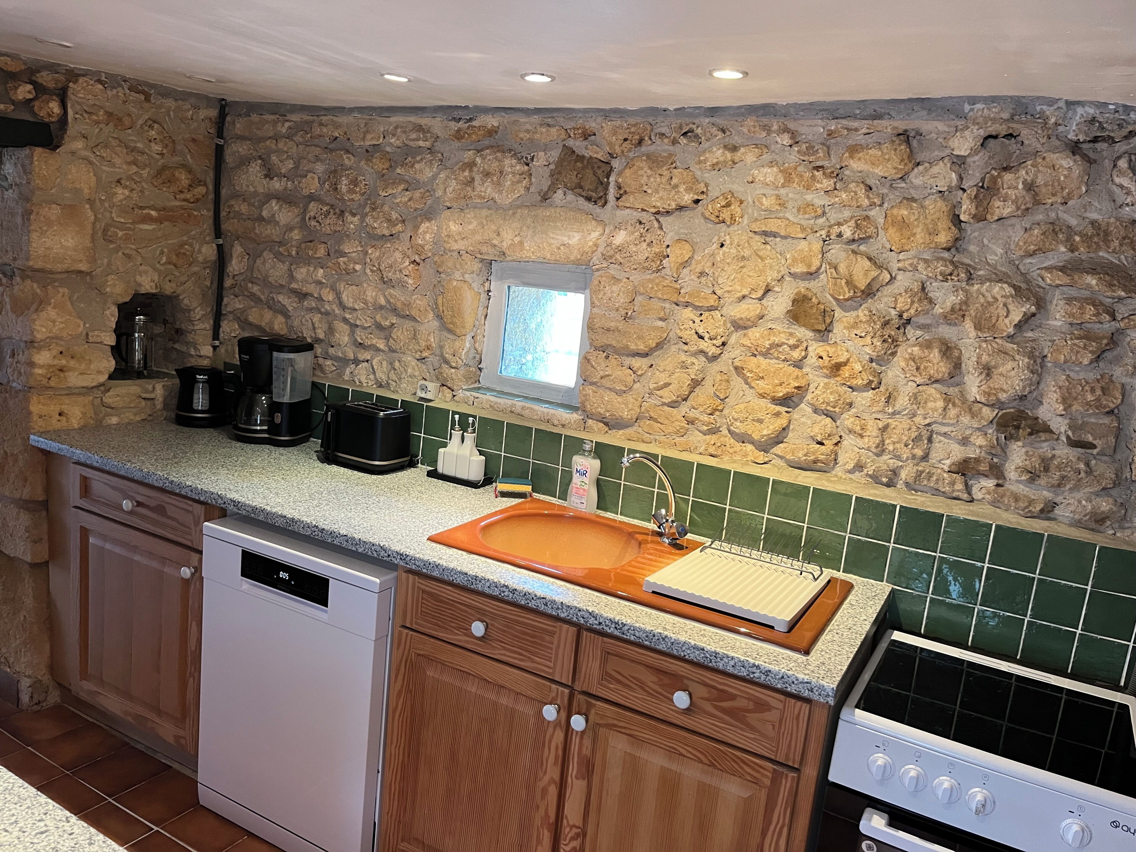 A kitchen with a stone wall, a window, a sink, a dishwasher, a stove, and wooden cabinets.
