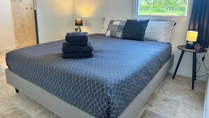Ein ordentlich bezogenes Bett mit einem geometrisch gemusterten Überwurf, ein Stapel gefalteter Handtücher, eine Nachttischlampe und ein kleiner Topfpflanze auf einem Beistelltisch.