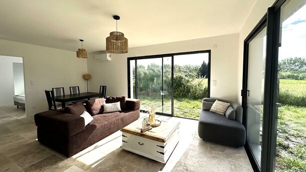 Ein modernes Wohnzimmer mit einem braunen Sofa, einem weißen Couchtisch und einem grauen Sessel. Es gibt einen Esstisch mit Stühlen und ein großes Fenster mit Blick auf Grünflächen.