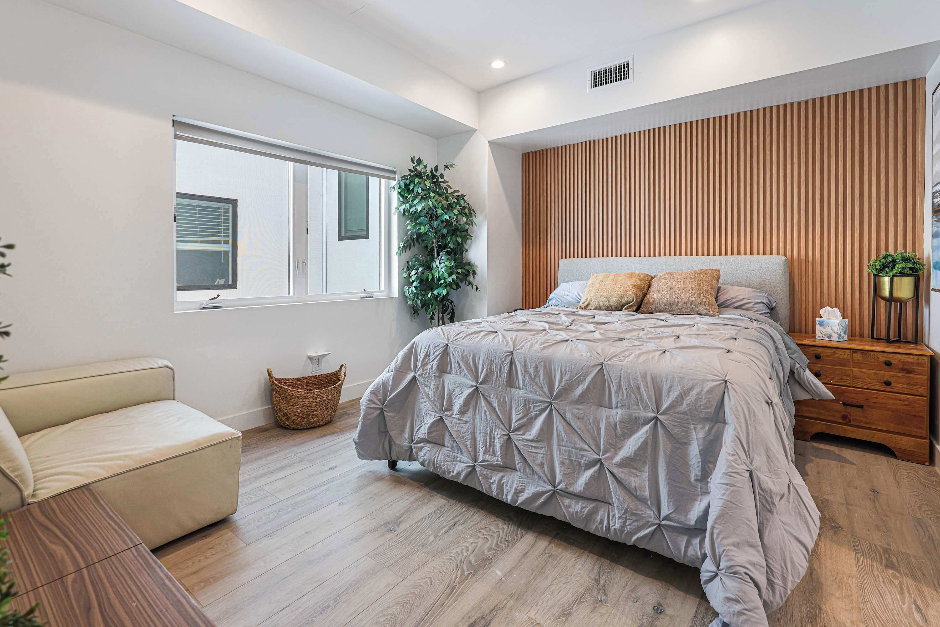 A modern bedroom with a bed, a wooden headboard, a window, a plant, and a bedside table.