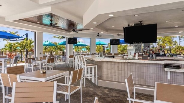 A spacious outdoor dining area with white chairs and tables, a bar with a tiled backsplash, and multiple screens mounted on the ceiling. - 2-bedroom apartment in delightful Venice with balcony, cool AC and gym pool! (Venice)