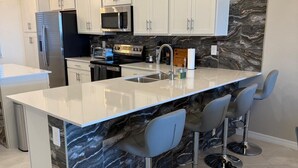 A modern kitchen with white cabinets, a marble backsplash, a stainless steel refrigerator, a microwave, an oven, and a sink. There are three bar stools with metal bases. - 2-bedroom apartment in delightful Venice with balcony, cool AC and gym pool! (Venice)