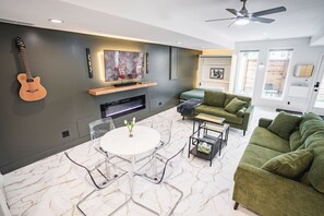 A modern living room with a fireplace, a television, a guitar, a green sofa, and a dining table with chairs. - Cozy KC Retreat, Private Suite w/ Patio &Fireplace (Kansas City)