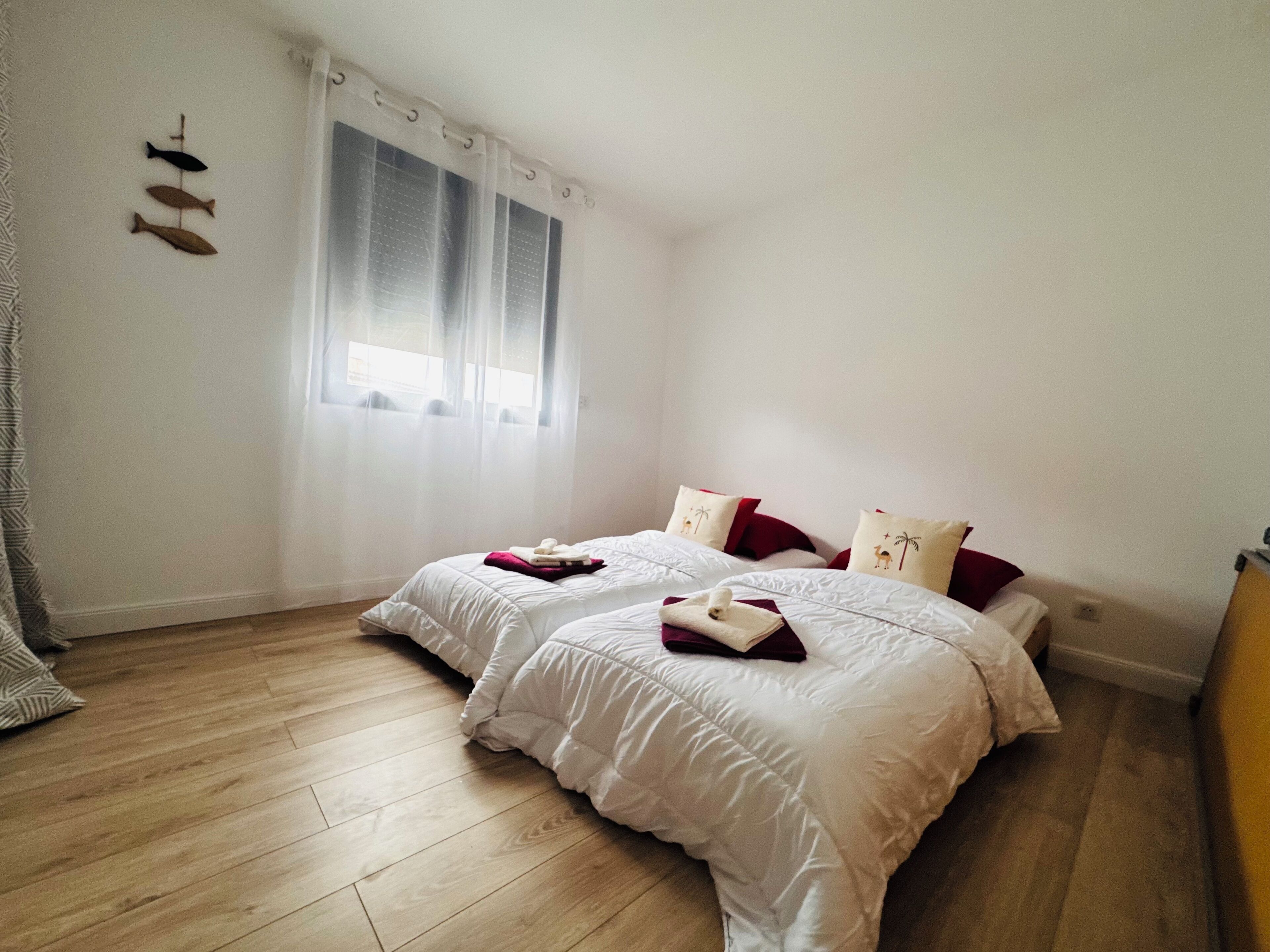 A bedroom with two beds, a window with sheer curtains, and a wooden floor.