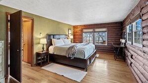 A log cabin bedroom with a bed, bedside tables, a desk, and a window. - Welcome to your peaceful 4 Br cabin, minutes from the historic Lake Junaluska. (Waynesville)
