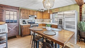 A kitchen with wooden cabinets, a stainless steel refrigerator, a chandelier, and a dining table set for four. - Welcome to your peaceful 4 Br cabin, minutes from the historic Lake Junaluska. (Waynesville)