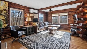 A log cabin interior with a sofa, armchair, and coffee table, large windows, and a ceiling fan. - Welcome to your peaceful 4 Br cabin, minutes from the historic Lake Junaluska. (Waynesville)