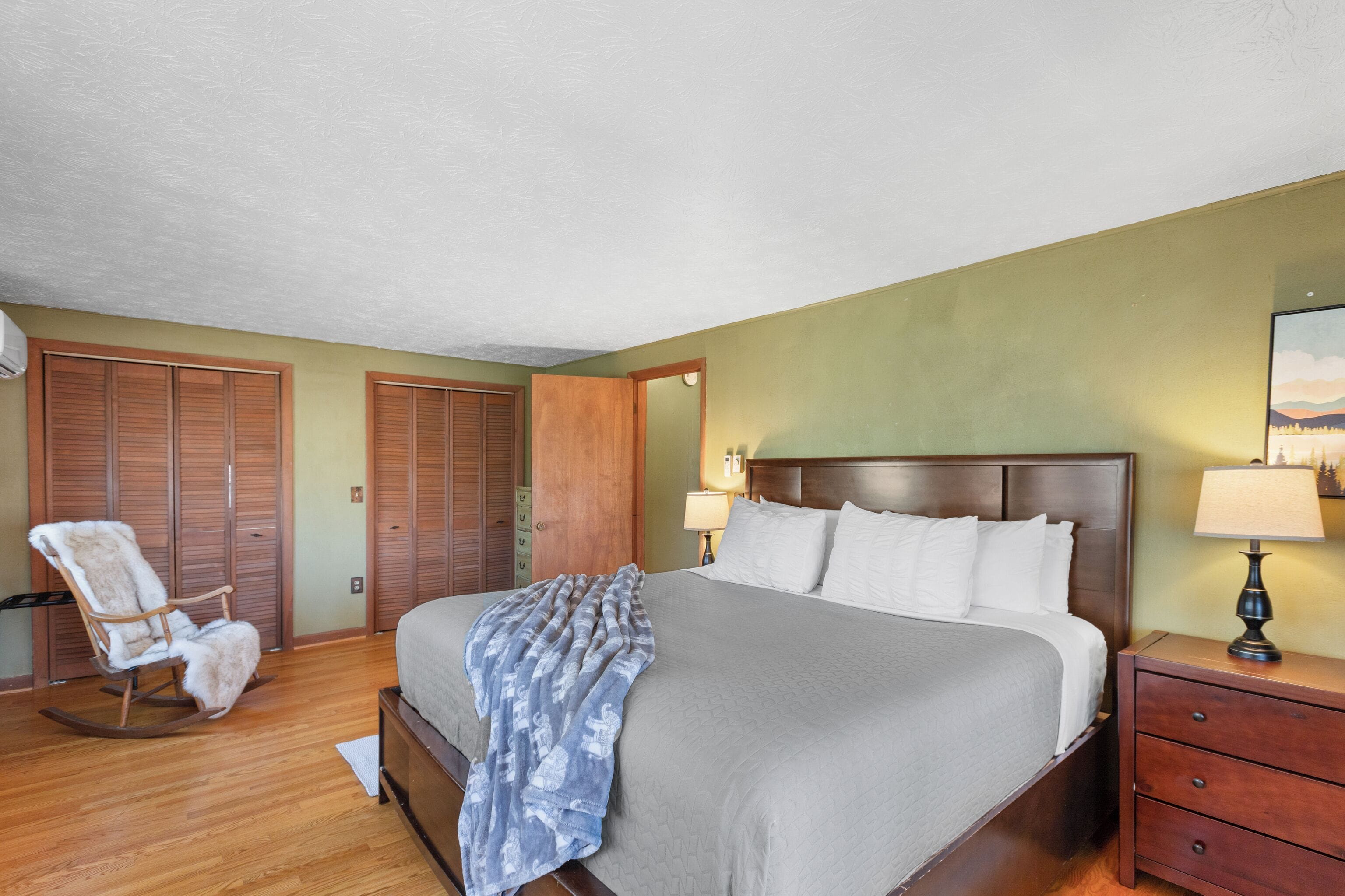 A bedroom with a bed, wooden wardrobe, rocking chair, bedside table, and lamp.